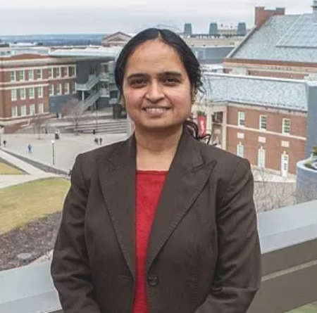 Shailaja Paik stands outside and faces the camera. She is smiling and wearing a dark brown blazer and red collarless shirt. She has a light brown skin tone and black hair.